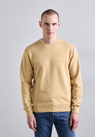 Sudadera color beige claro con cuello redondo, mangas largas y puños acanalados. En la parte frontal se muestra el texto "TIGER OF SWEDEN" en blanco.