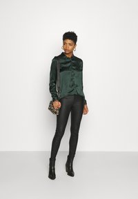 Blusa di satin verde smeraldo con bottoni frontali, abbinata a pantaloni in ecopelle neri e stivaletti neri. Borsa con stampa leopardata come accento.