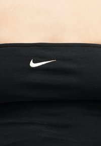 Μαύρο ύφασμα με λευκό λογότυπο Nike swoosh κεντημένο στο κέντρο ενός ρούχου χωρίς τιράντες πάνω σε ανοιχτόχρωμο δέρμα.