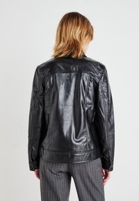 Sort læderjakke med en glat, blank overflade, der har en klassisk krave og struktureret rygdesign. Båret med stribede bukser.