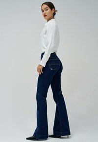 Jeans de mezclilla acampanados en índigo oscuro, de cintura alta con un diseño clásico de cinco bolsillos. Combinados con una blusa blanca de manga larga y sencilla.