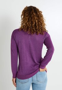 Pull violet à manches longues avec une finition texturée et scintillante. Doté d'un ourlet arrondi et de poignets côtelés, associé à un jean bleu clair.