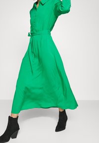 Grünes Langarm-Hemdkleid aus glattem Stoff mit einem taillierten Gürtel, fließendem Rock und schwarzen Stiefeletten mit Blockabsatz.