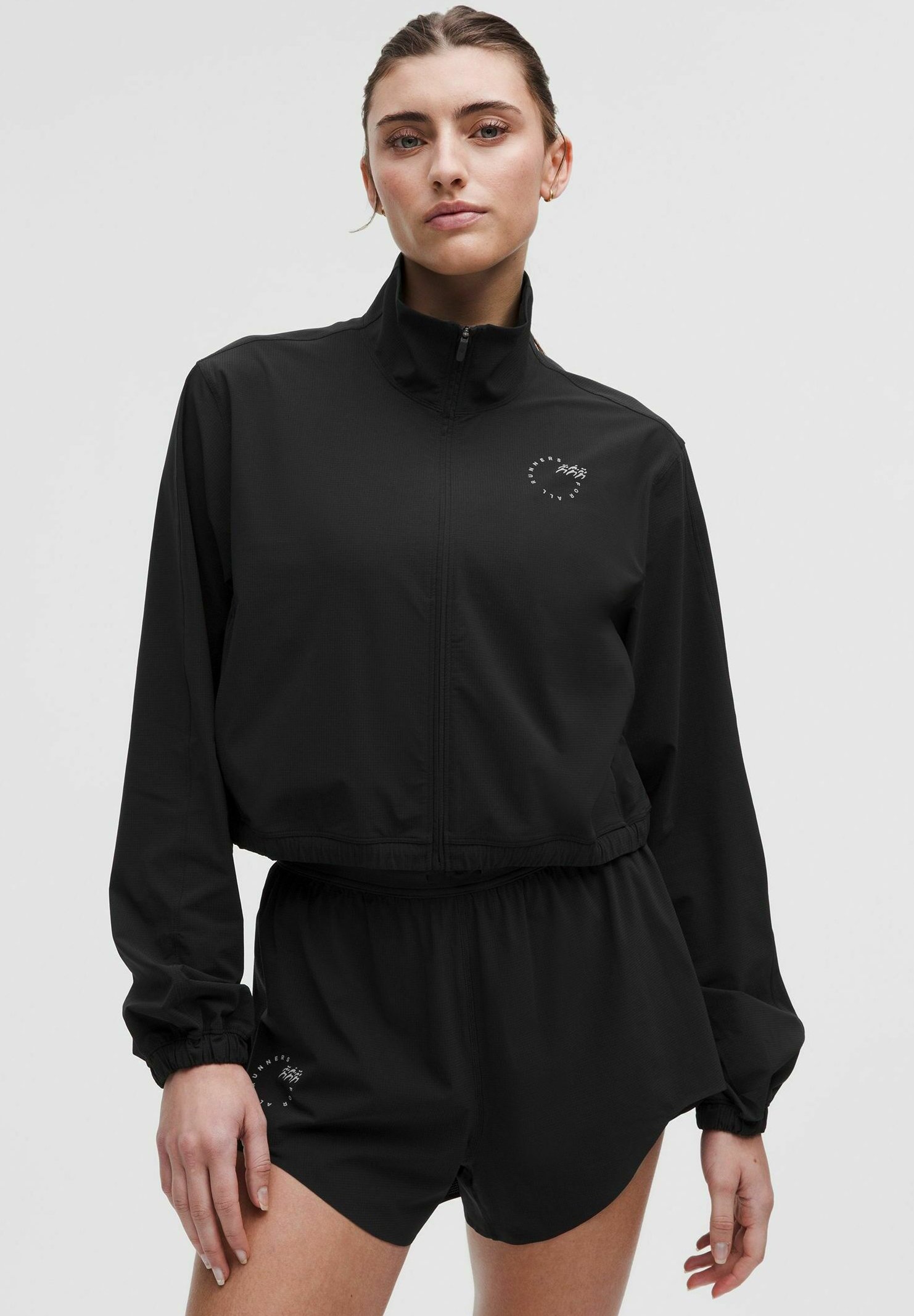 Črna športna jakna z zadrgo, visokim ovratnikom in elastičnimi manšetami, v kombinaciji s kratkimi hlačami, ki imajo logotip na strani. Gladek material.