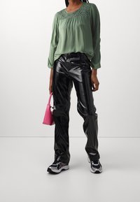 Grön blus med spetsdetaljer, svarta glänsande vida byxor, svartvita sneakers och en liten rosa handväska.