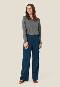 Grijze lange mouwen top, losse pasvorm, gecombineerd met donker teal wijde cargo broek met zijzakken, aangevuld met bruine sandalen.