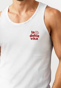 Weißes ärmelloses Tanktop mit einem roten Grafikdruck, der "la dolce vita" mit einem Blumendetail auf der linken Seite zeigt.