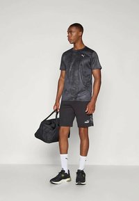 Mustas Puma sportlikus riietuses mees hoiab musta spordikotti, tal on jalas valged sokid ja mustad tossud, seistes lihtsa tausta ees.