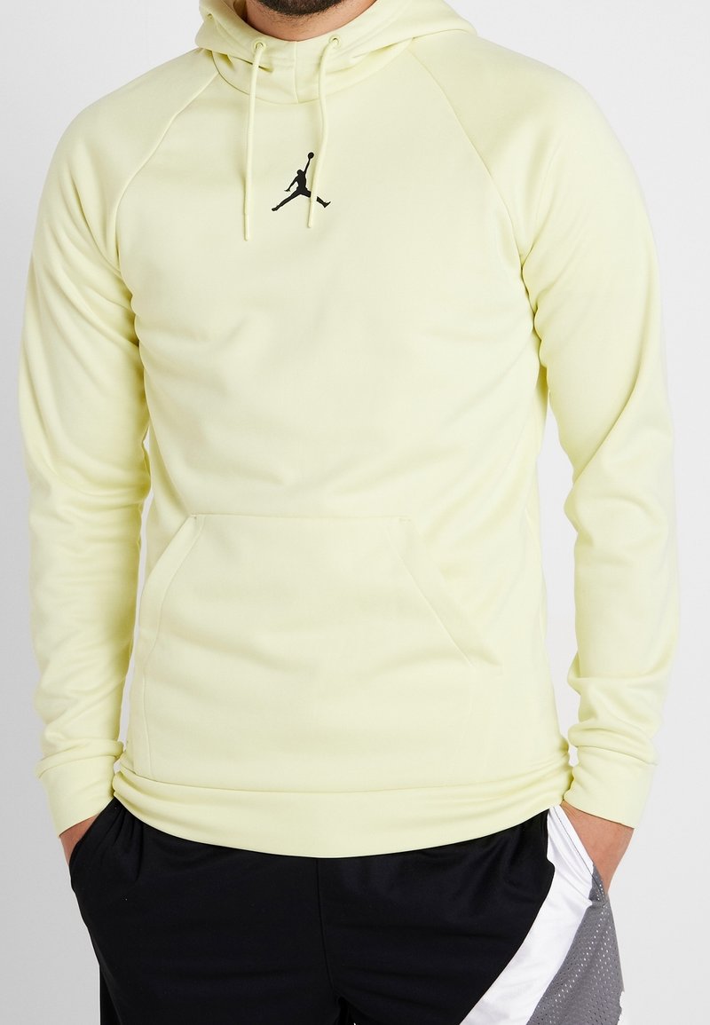 Homme portant un sweat à capuche jaune pâle avec un logo Air Jordan noir et un short de sport noir et blanc, mains dans les poches.