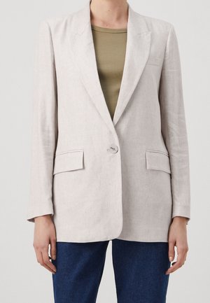 Blazer beige ajusté avec un bouton unique, deux poches à rabat à l'avant, porté sur un haut côtelé olive et un jean bleu.