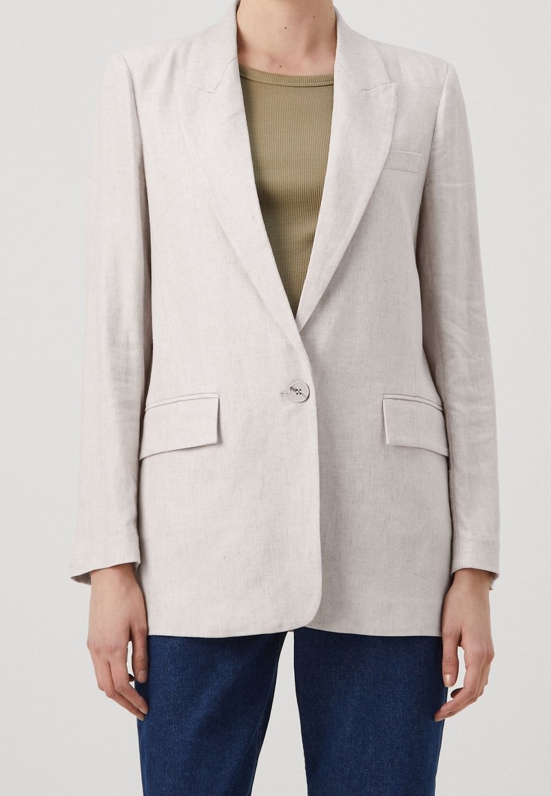 Blazer beige entallado con un solo botón, dos bolsillos delanteros con solapa, llevado sobre una camiseta de canalé verde oliva y vaqueros azules.