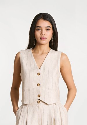 Jeune femme aux longs cheveux foncés portant un gilet sans manches beige à rayures et un pantalon plissé assorti, debout les mains dans les poches.