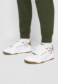 Vita sneakers med beige detaljer, texturerade överdrag och en låg design. I kombination med olivgröna joggers med cuffade nederdelar.