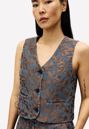 Femme portant un gilet sans manches marron avec un motif floral bleu et un pantalon assorti, montrant des tatouages sur les deux bras contre un fond uni.