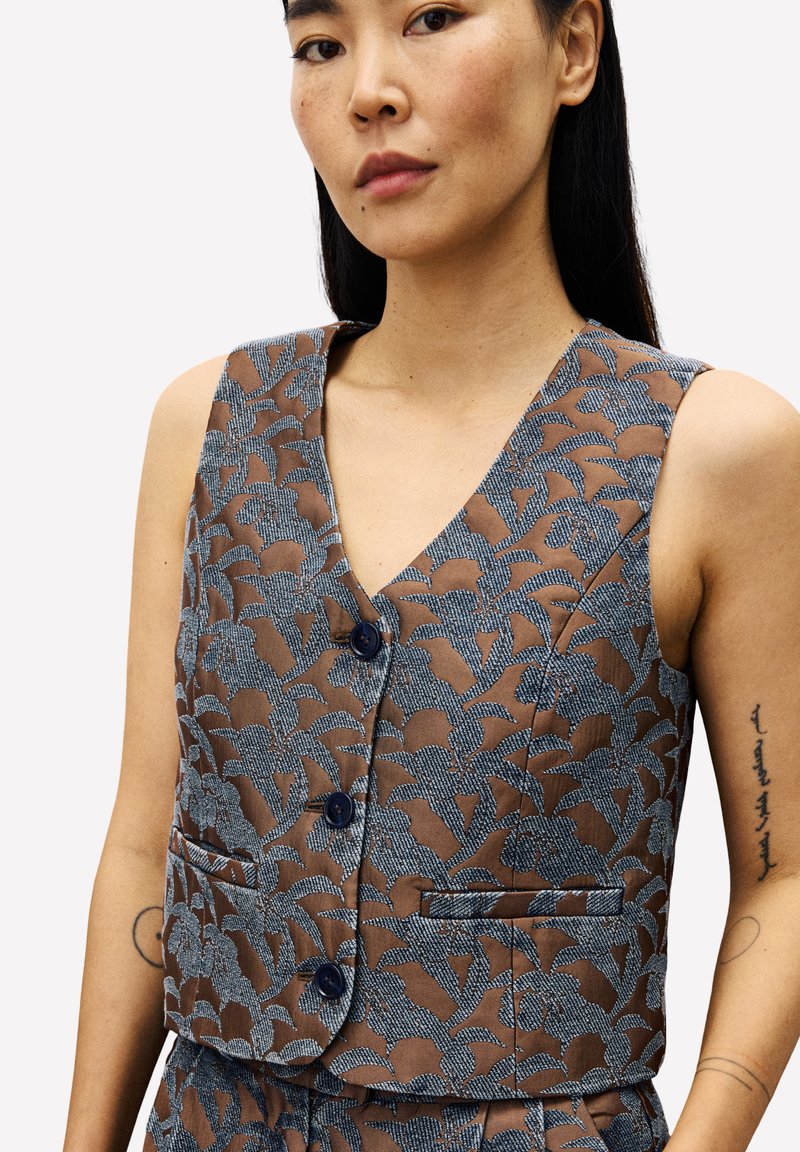 Vrouw in een mouwloze bruine vest met een blauw bloemenpatroon en bijpassende broek, met tattoos op beide armen tegen een effen achtergrond.