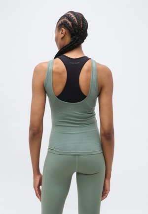 Γυναίκα με πλεξούδες, που φοράει μαύρο αθλητικό σουτιέν, πράσινο αμάνικο τοπ και κολάν, εμφανίζεται από πίσω μπροστά σε απλό φόντο.