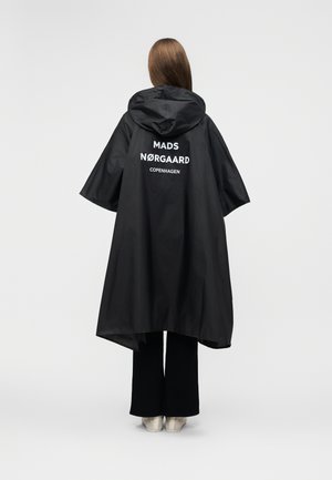 Άτομο που φοράει μαύρο αδιάβροχο με κουκούλα, με το κείμενο "MADS NØRGAARD COPENHAGEN" στην πλάτη, συνδυασμένο με μαύρο παντελόνι και λευκά παπούτσια.