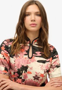 Top con estampado floral que presenta un cuello alto, mangas cortas y un detalle de ojal. Los colores incluyen negro, rosa y crema con un acabado texturizado.