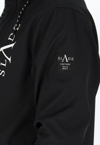 Schwarze Sportjacke aus glattem, elastischem Material. Verfügt über ein weißes Logo und ein rechteckiges Markenlabel am Ärmel.