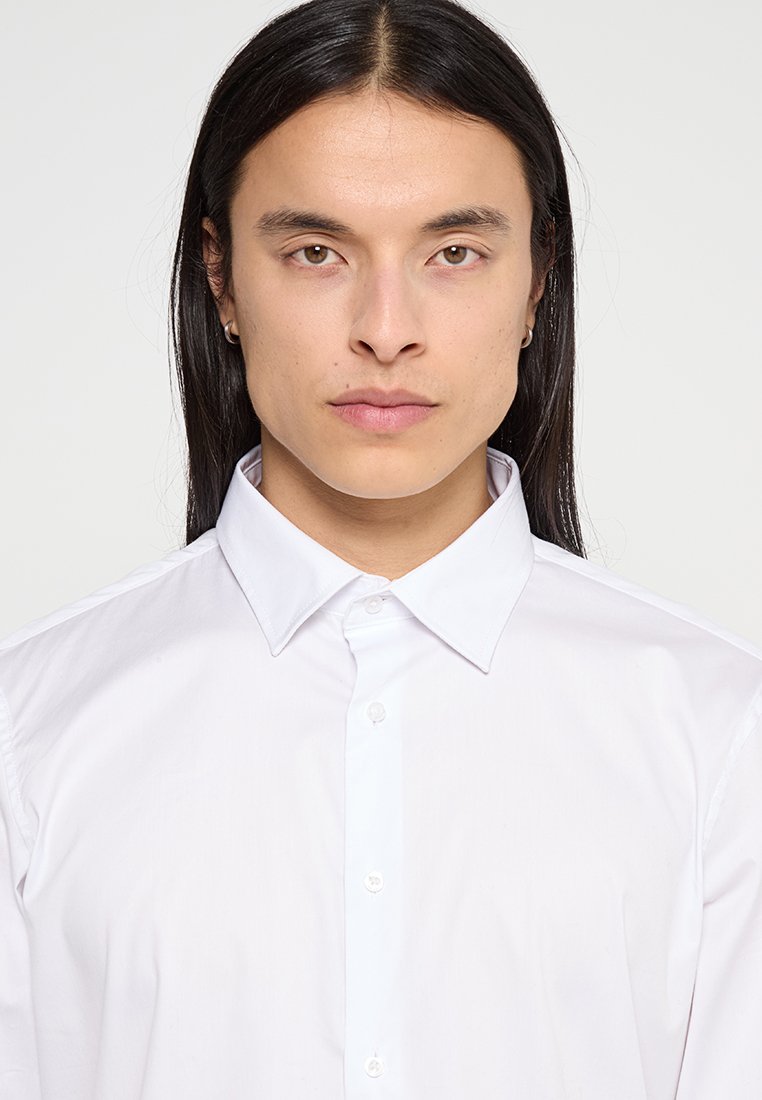 Jeune personne avec de longs cheveux noirs, portant de petites boucles d'oreilles en anneau et une chemise blanche boutonnée, tournant le visage vers l'avant avec une expression neutre.