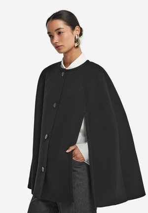 Cape en laine noire au design arrondi, dotée de trois boutons et de fentes latérales. Portée sur une chemise blanche à col, sur fond uni.