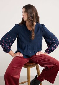 Blusa blu navy con maniche a sbuffo, chiusura con bottoni, ricamo floreale rosa sui polsini, dettaglio plissettato, abbinata a pantaloni larghi in corduroy rossi.