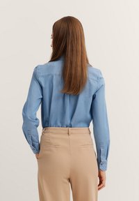 Himmelblaues Langarmhemd mit Kragen, geknöpften Manschetten und Rückenpasse, kombiniert mit beigen Hosen, die eine glatte Textur und seitliche Taschen aufweisen.