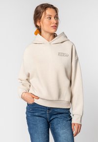 Beige hoodie met een zachte textuur, oversized pasvorm en kap. Voorzien van een klein gedrukt tekstdetail op de voorkant. Gecombineerd met blauwe jeans.