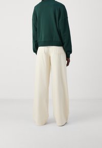 Grüner, verkürzter Pullover mit tiefen Schultern, kombiniert mit weiten cremefarbenen Hosen mit Gesäßtaschen und einer glatten, matten Textur.