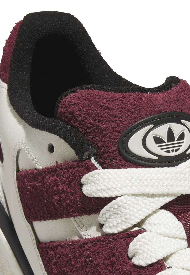 Sneaker blanc avec des accents en daim bordeaux, comportant un col noir, des lacets blancs et un logo ovale avec trois rayures sur le côté.