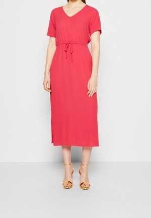 Femme portant une robe corail mi-longue à manches courtes, encolure en V, taille à cordon, et sandales dorées à talons hauts, debout sur un fond uni.
