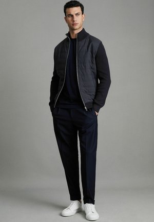 Dunkel gesteppte Jacke mit gerippten Ärmeln, marinefarbene Unterlage, dunkle Hose und weiße Sneaker. Das Modell posiert mit den Händen in den Taschen vor einem neutralen Hintergrund.