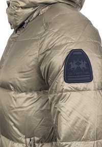 Olivově zelená puffa bunda s prošívaným designem, s námořnickým modrým logem na levém rameni a předním zipem.