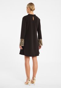 Schwarzes Kleid mit langen Ärmeln und strukturierten Leopardenmuster-Akzenten. Rückenausschnitt mit Schlüsselverschluss und ausgestellter Silhouette. Goldene Absatzschuhe.