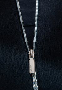 Navy Fleecejacke mit Reißverschluss im Close-up, ausgestattet mit einem silbernen Metallreißverschluss und strukturiertem Stoff. Der Reißverschlussgriff ist mit einer Markengravur versehen.