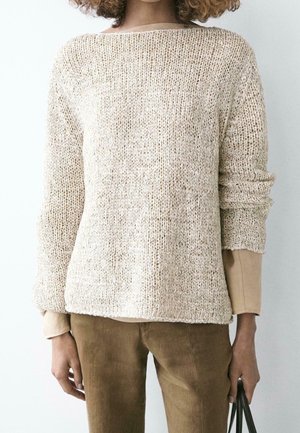Personne portant un pull en maille crème ample par-dessus un haut beige à manches longues et un pantalon marron, tenant un sac noir dans une main.