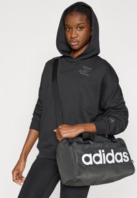 Crna majica s kapuljačom i detaljem logotipa sprijeda, uparena s crnom torbom za putovanje s velikim bijelim natpisom "adidas". Glatka tekstura, sportski dizajn.