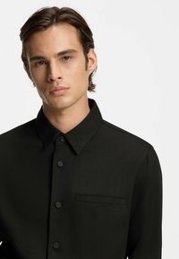 Chemise noire à boutons avec un col structuré, des manches longues et une poche poitrine gauche. Le tissu semble lisse avec une texture subtile.
