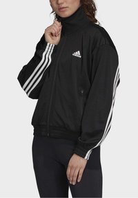 Black zip-up jacket with high collar, featuring white three-stripe accents on sleeves and elastic cuffs. Material appears smooth and durable.