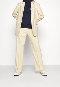 Beige kavaj över en marinblå huvtröja, parat med krämfärgade vida byxor och mönstrade sneakers. Slät tyg med strukturerad design.