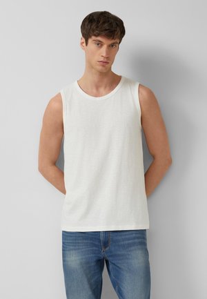 Mężczyzna ubrany w biały top bez rękawów i niebieskie dżinsy, stoi oparty o jasnoszarą ścianę z rękami za plecami.