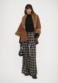 Mujer vestida con una capa de piel marrón con ribete, un suéter de cuello alto negro, pantalones de cuadros de pierna ancha, zapatos negros y sosteniendo un bolso negro, de pie sobre un fondo blanco.