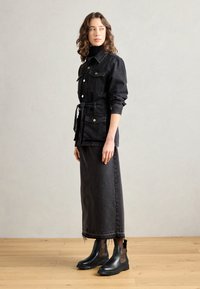 Giacca di jeans nera con tasche, vita imbottita e chiusura con bottoni, abbinata a una gonna lunga coordinata e stivaletti neri con dettagli leopardati.