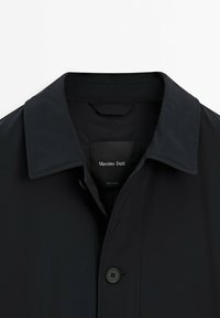 Schwarzer wasserdichter Trenchcoat mit klassischem Kragen, Frontknopfverschluss und einem Etikett mit "Massimo Dutti". Glatte Textur.