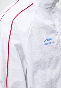 Λευκό αδιάβροχο μπουφάν με κόκκινη ρίγα στον ώμο και μπλε λογότυπο ARKK στο στήθος, με το μπροστινό φερμουάρ μερικώς ορατό.