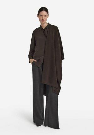 Donna che indossa una camicia marrone scuro con bottoni, pantaloni ampi grigio scuro e uno scialle drappeggiato marrone scuro, in piedi contro uno sfondo bianco.