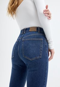 Jeans de mezclilla azul oscuro con una cintura alta, que presentan bolsillos traseros y costuras en contraste. Etiqueta de la marca en tono marrón en la cintura. Textura suave.