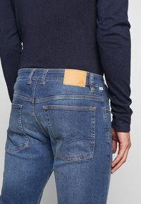 Modré džíny z denimu s úzkým střihem, s koženou nášivkou v barvě tan na pase, zadními kapsami a jemným vyblednutím na látce.