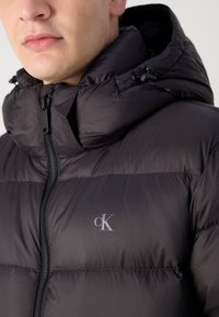 Black puffer jacket with a high collar, zip closure, and a small CK logo on the chest. Features a smooth, shiny texture and adjustable hood.