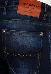 Dunkelblaue Jeans aus Denim mit orangefarbenen Nähten und einem braunen Lederetikett. Zu den Merkmalen gehören eine Gesäßtasche mit einem dekorativen Emblem und einer Niet.